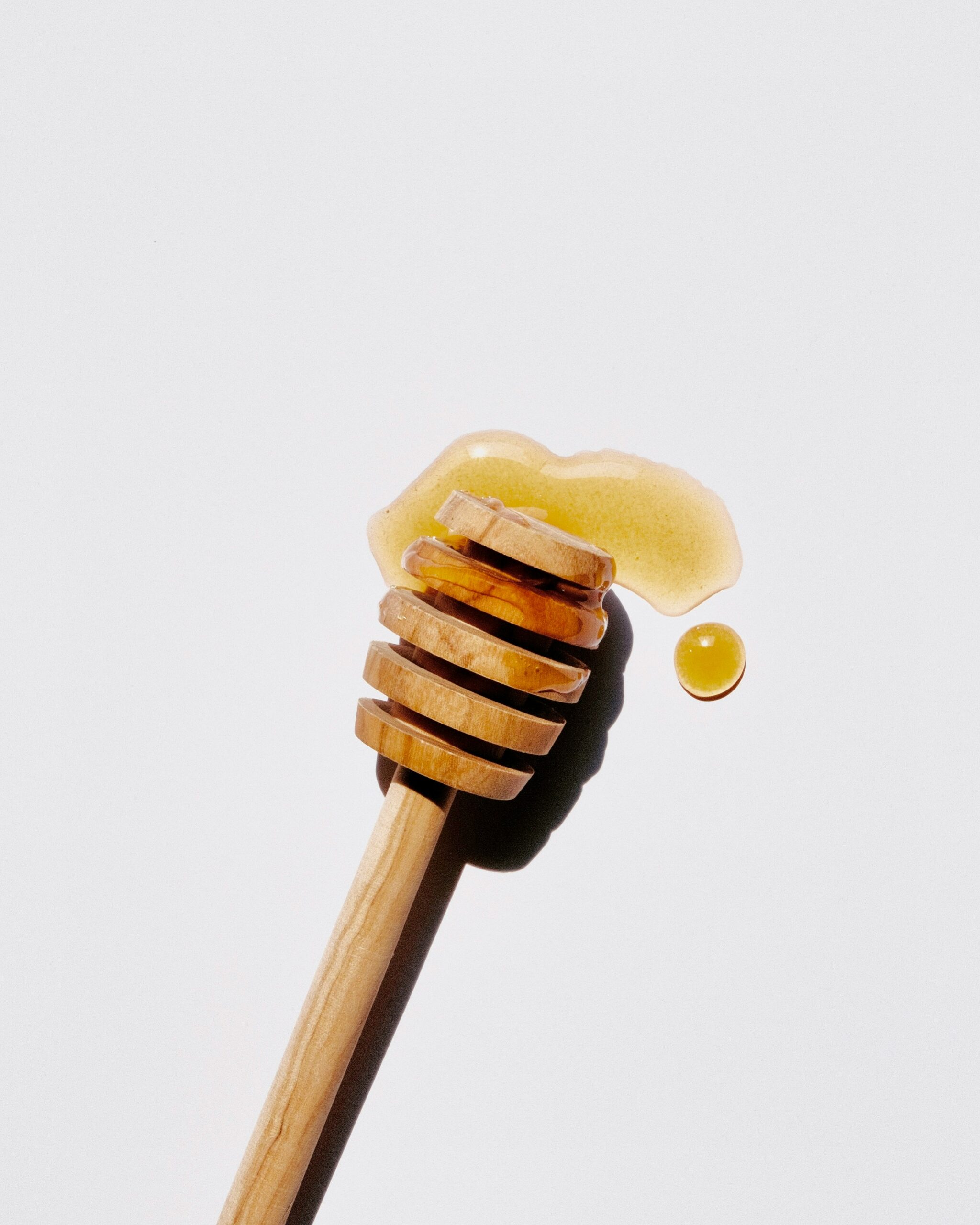 A honey dipper with honey on it and a small drop of honey beside it, placed on a plain white background.