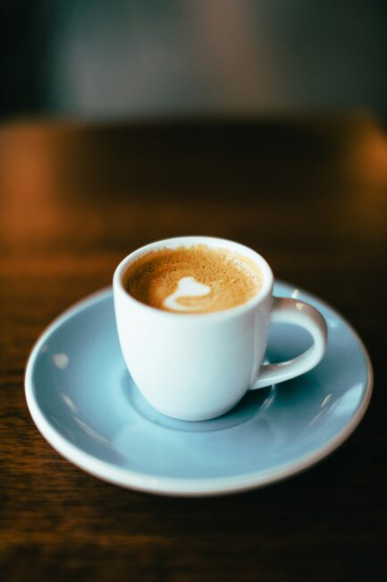 A white ceramic cup of coffee with foam sits on a matching saucer on a wooden table.