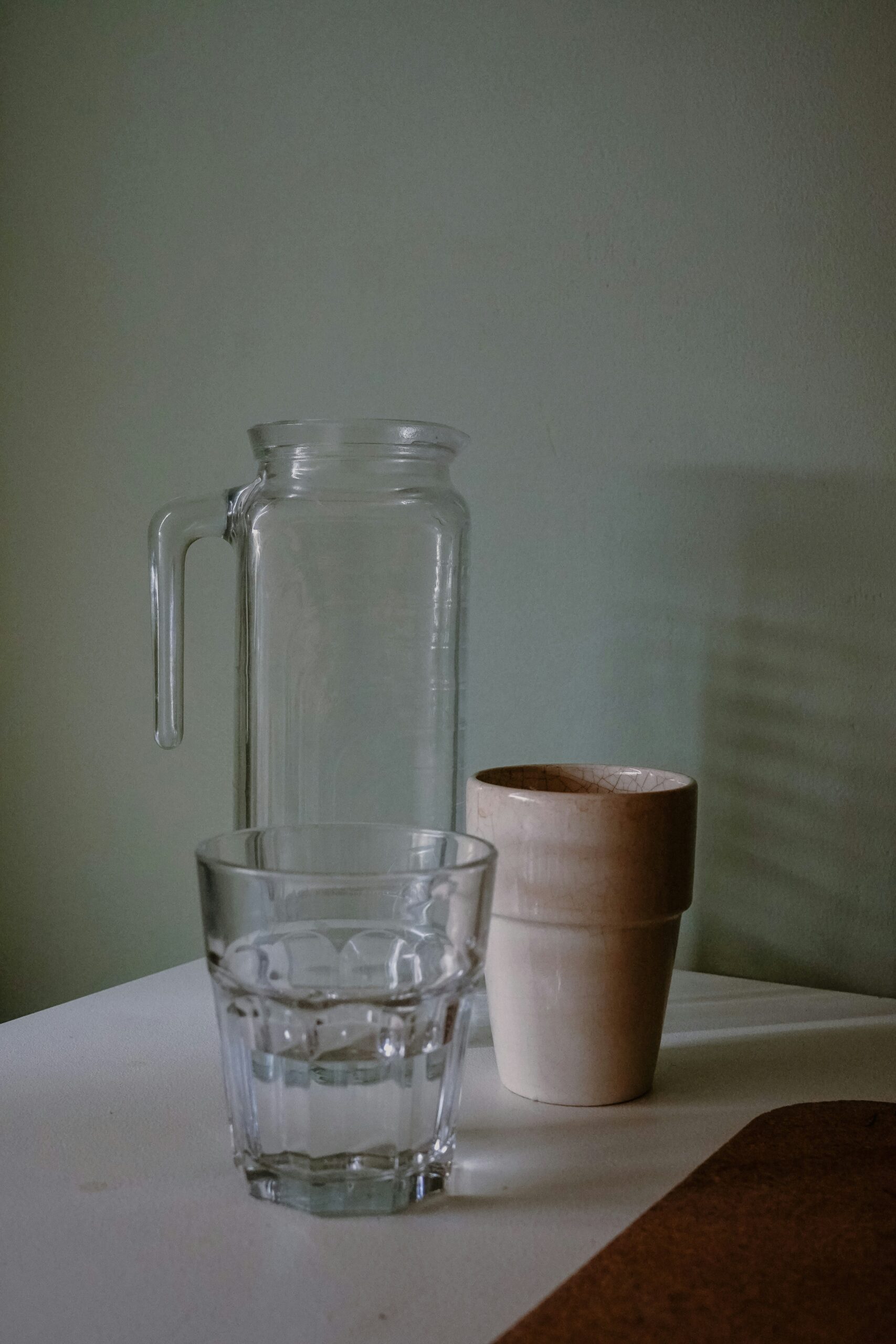 A clear glass jug, a glass of water, and two stacked ceramic cups are arranged on a white surface against a plain light green wall.