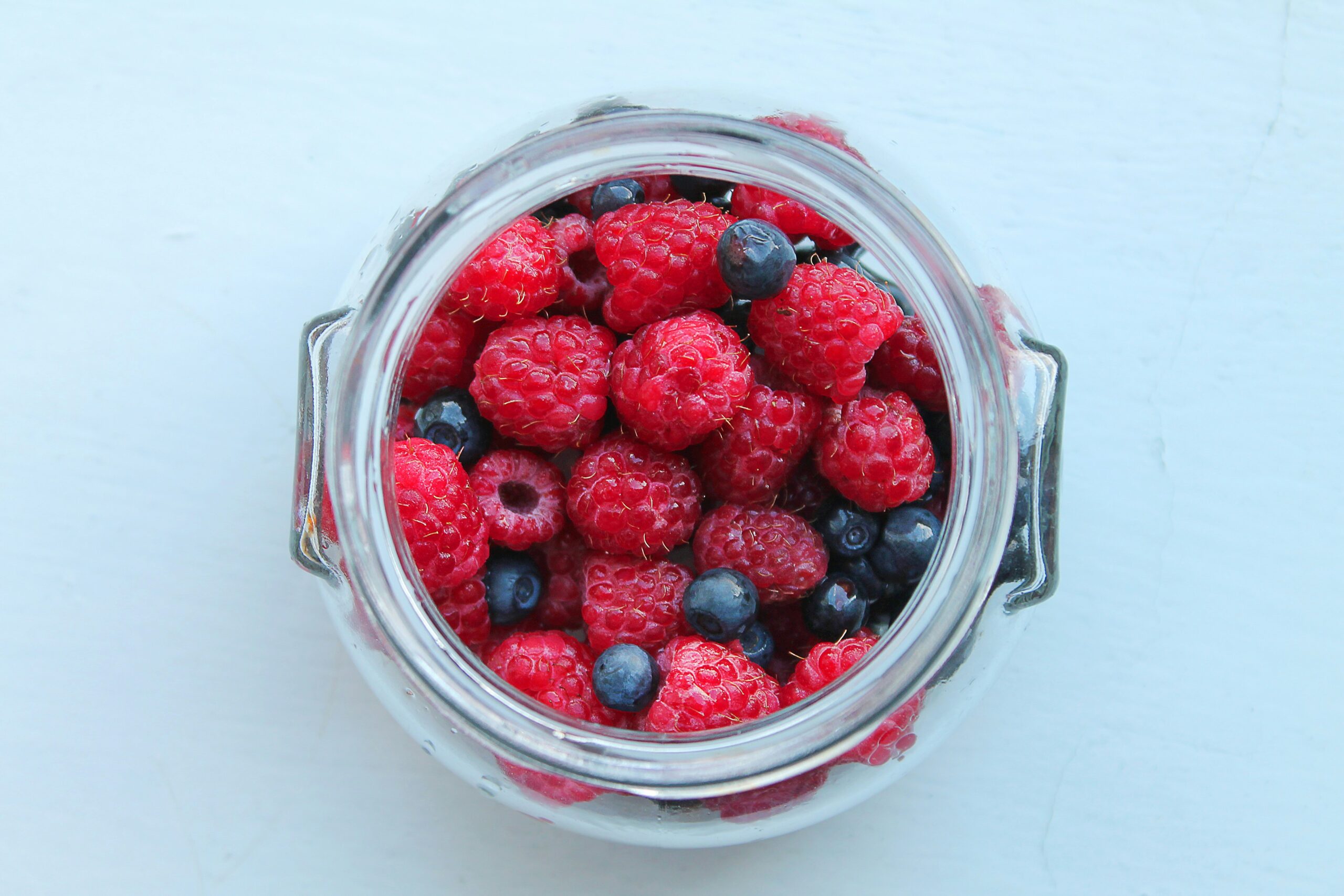 A glass jar filled with fresh raspberries and blueberries viewed from above, set against a light background.