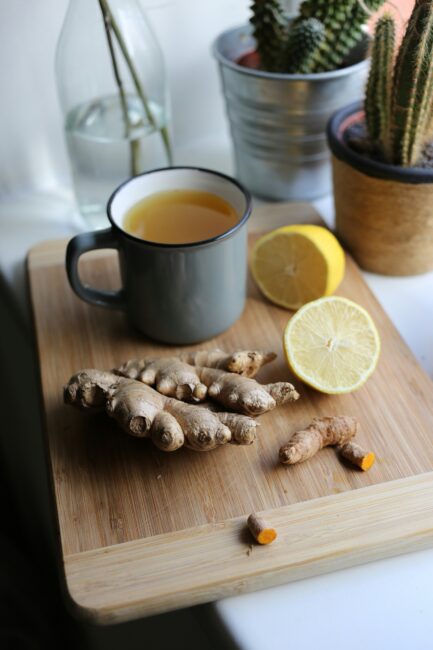 A mug of tea, fresh ginger, turmeric root, and a halved lemon are arranged on a wooden cutting board near potted cacti and a glass bottle.