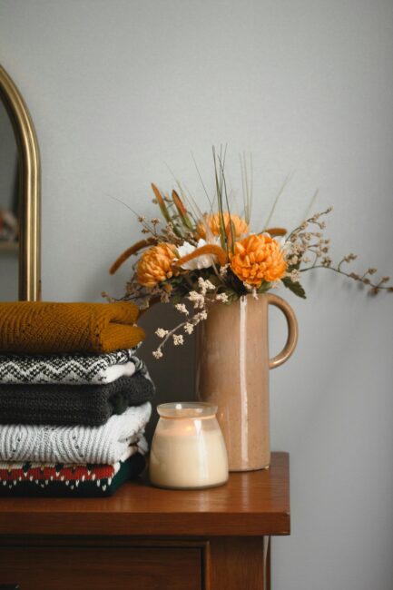 A stack of folded sweaters, a lit candle, and a beige vase with orange flowers sit on a wooden dresser against a gray wall.