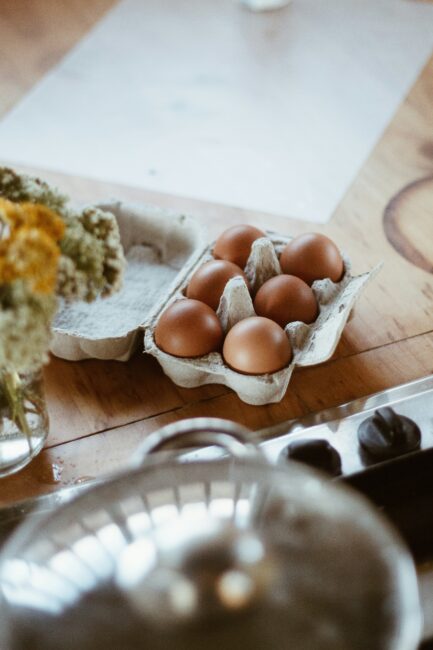 A half-dozen brown eggs in an open carton sit on a wooden table near a vase of flowers and a reflective metal lid.