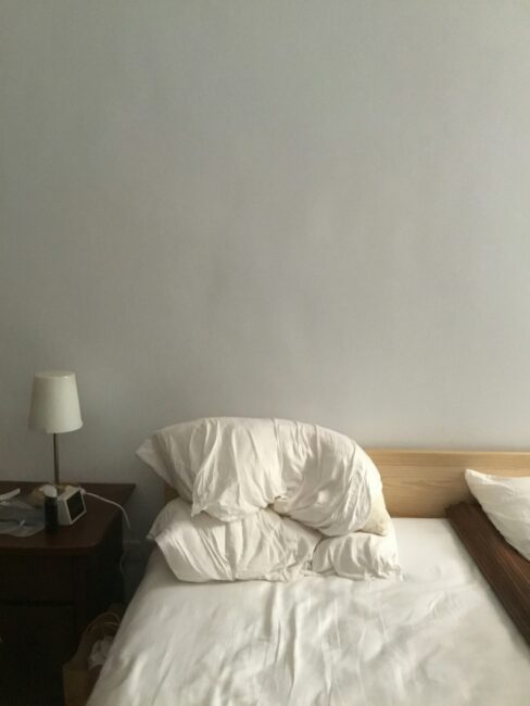 A neatly made bed with rumpled white pillows and sheets, next to a wooden nightstand with a lamp, a clock, and a few small items, against a plain white wall.