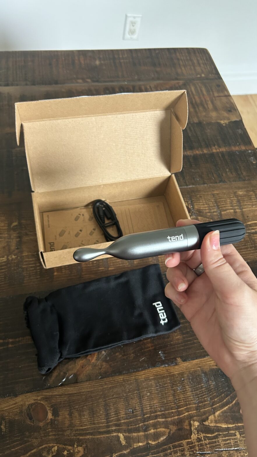 A hand holding a Tend electric toothbrush above its open box, with a charging cable and a black carrying pouch on a wooden table.