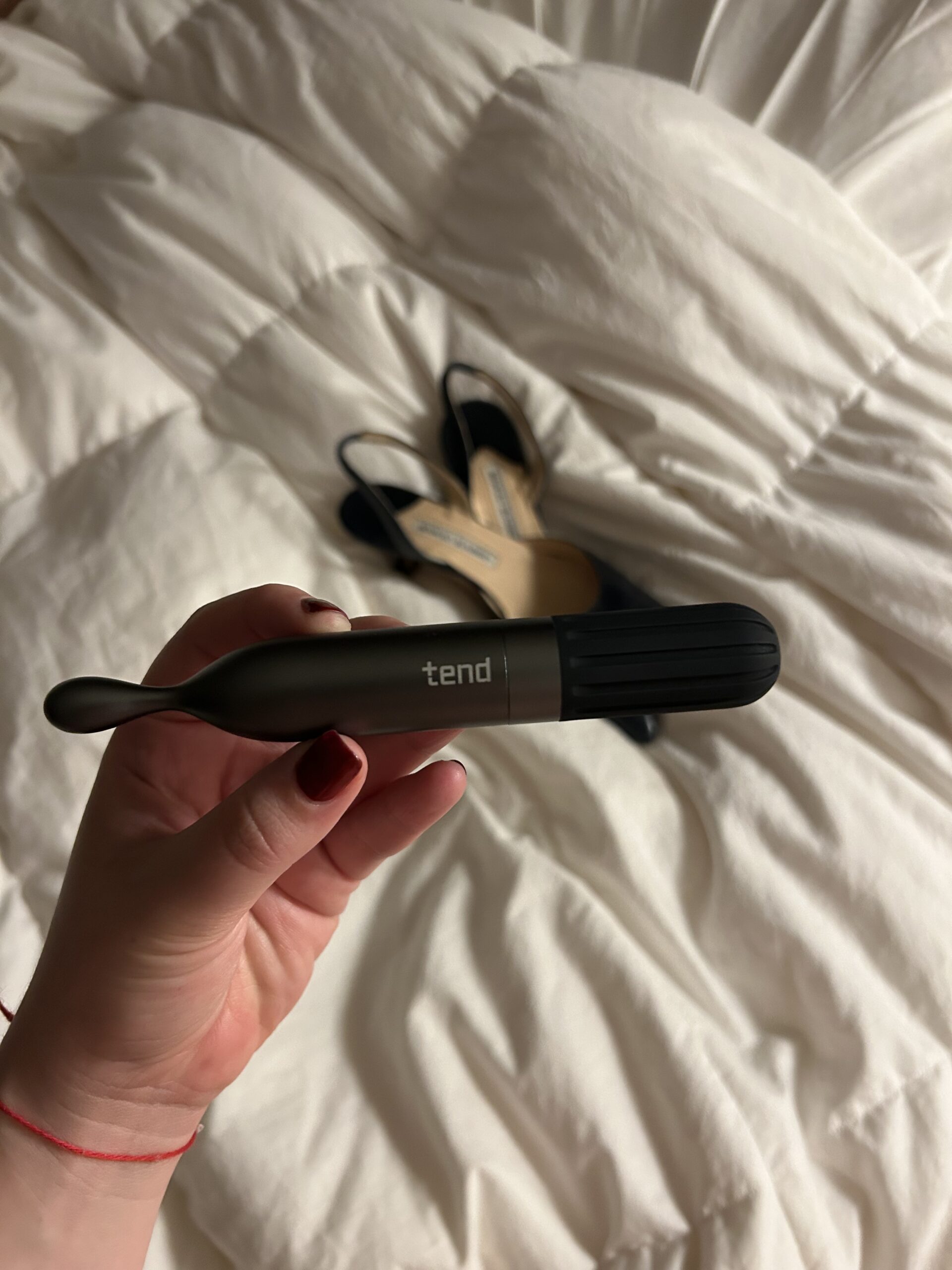 A hand holds a black and gray Tend toothbrush with a curved handle; a pair of black heeled shoes is on a white, rumpled bed in the background.