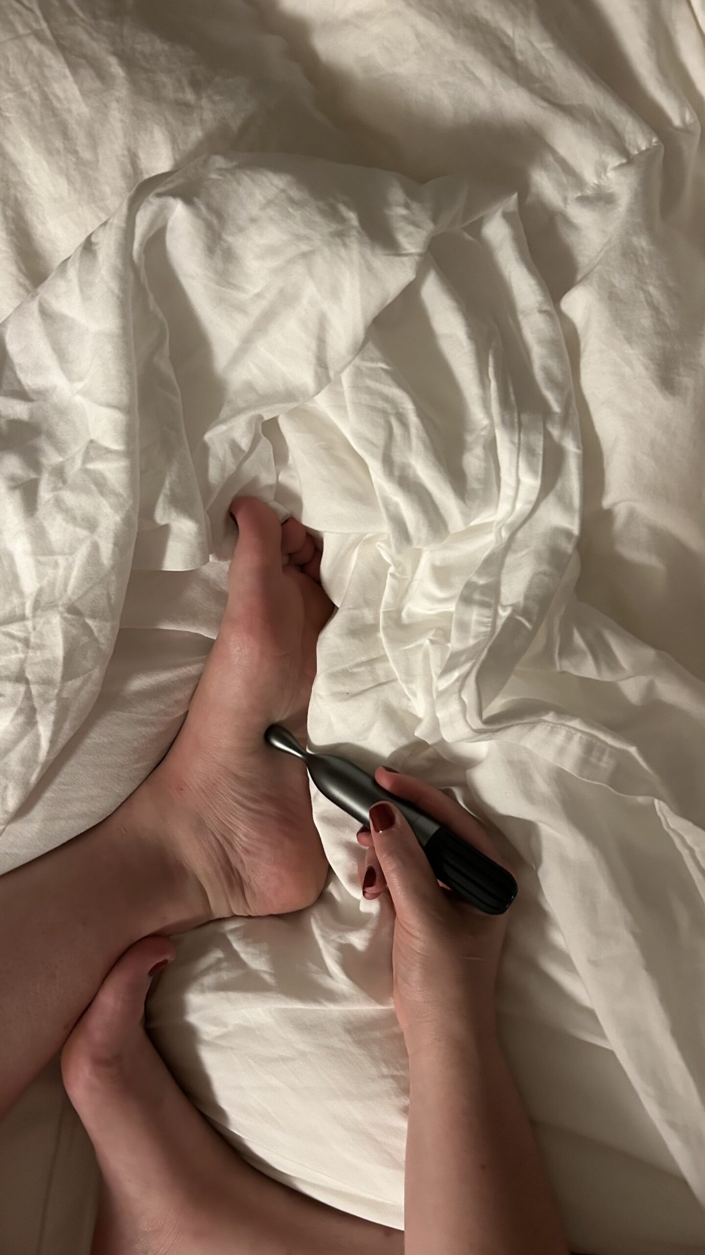 A person uses a handheld massage tool on the sole of their foot while lying on a white bed.