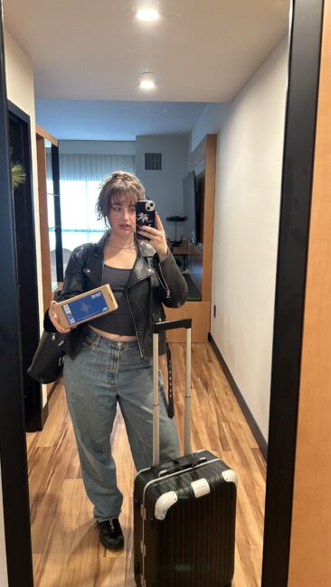 Person taking a mirror selfie in a room, holding a box of Tend, with a suitcase and shoulder bag, wearing a leather jacket, crop top, and jeans.