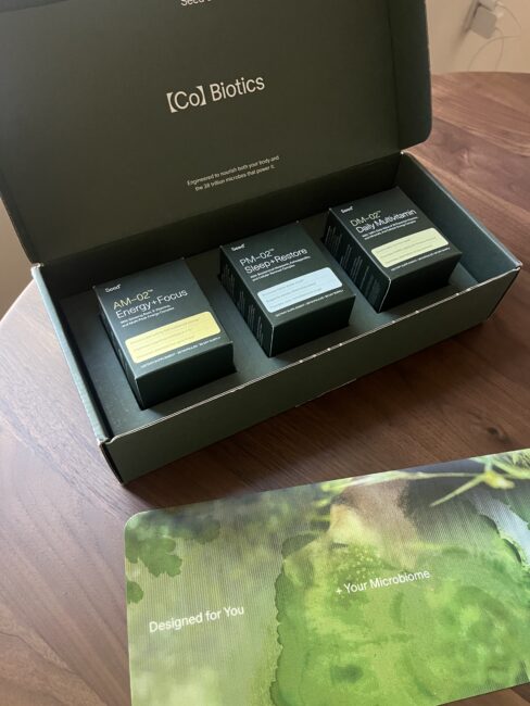 An open Biotics box on a wooden table contains three supplement packages labeled AM-02 Energy+Focus, PM-01 Stress+Relaxing, and Daily-01 Gut+Wellbeing. A pamphlet is also visible.