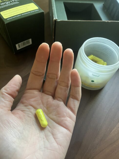 A hand holds a yellow capsule above a wooden surface, next to an open jar containing similar capsules and a green box.