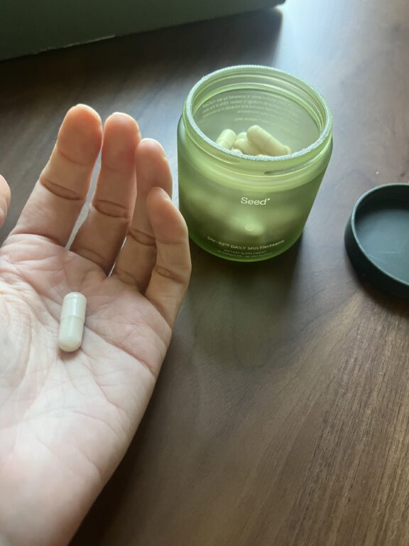 A hand holds a white capsule next to an open green jar labeled "Seed" containing more capsules, on a wooden surface.