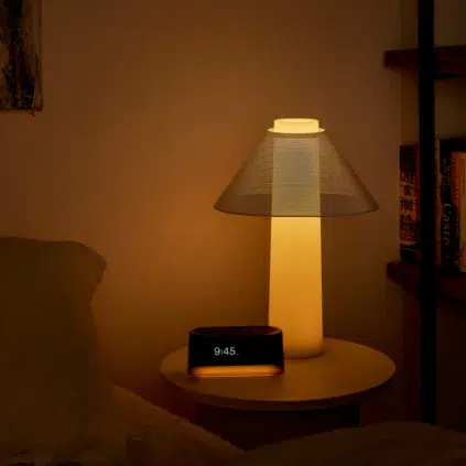 A softly glowing lamp, a digital clock showing 9:45, and a couple of massage guns rest on a round bedside table next to a bed and a bookshelf.