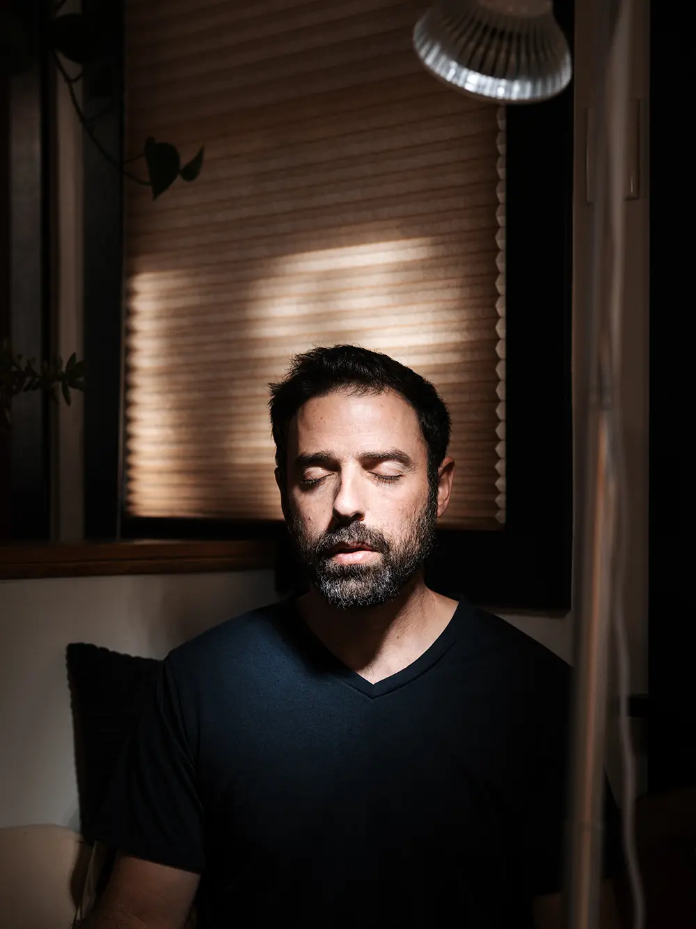 A man with a beard and closed eyes sits indoors in soft lighting, wearing a dark t-shirt, with window blinds in the background, enjoying the soothing effects of massage guns.
