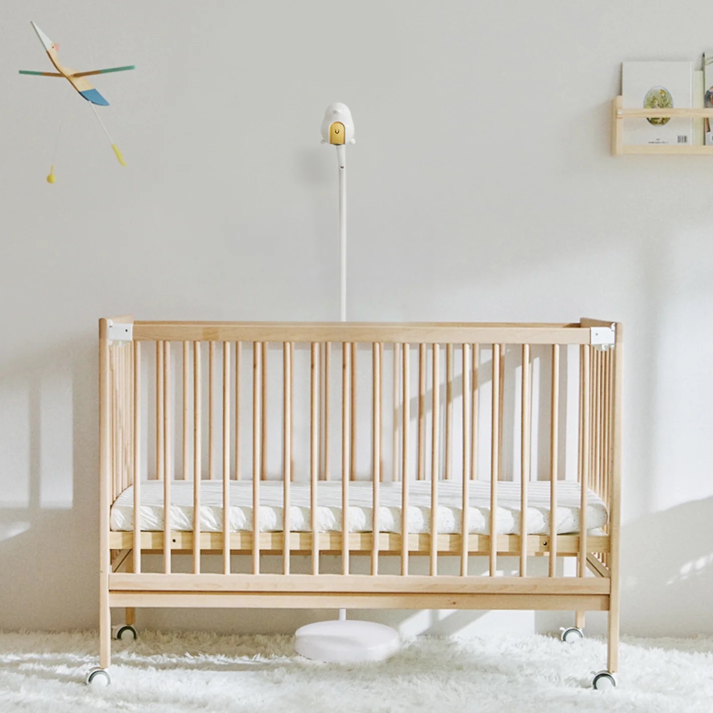A wooden baby crib with a mattress sits on a white rug. A floor lamp, wall shelf, massage guns, and mobile add subtle touches to the minimal, bright room.