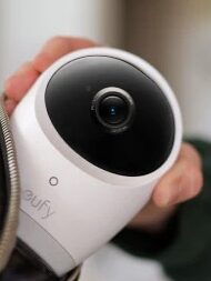 A person places a Eufy security camera into a black bag, with the camera lens facing outward, alongside other gadgets like massage guns for on-the-go convenience.