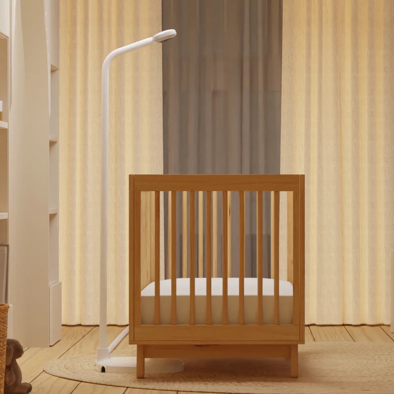A wooden crib with a white mattress stands on a wooden floor next to a white floor lamp and massage guns, in front of beige curtains in a softly lit room.