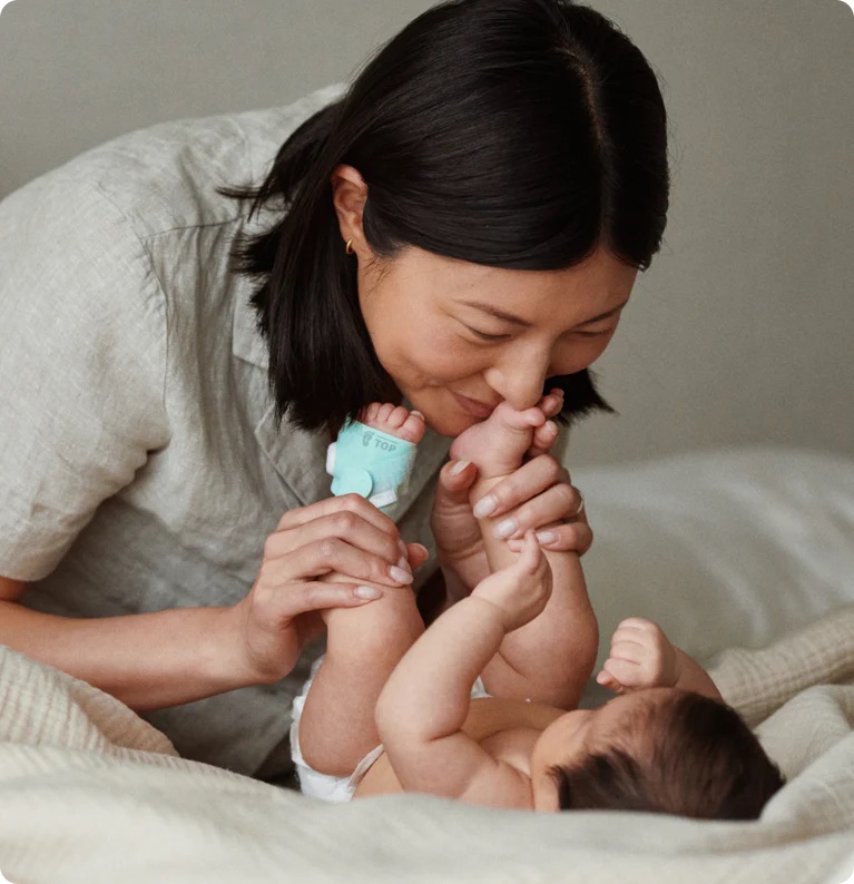 An adult gently holds and kisses a baby's feet while the baby lies on a bed, wearing a small teal monitor on its foot—capturing tender moments far removed from the intensity of massage guns.