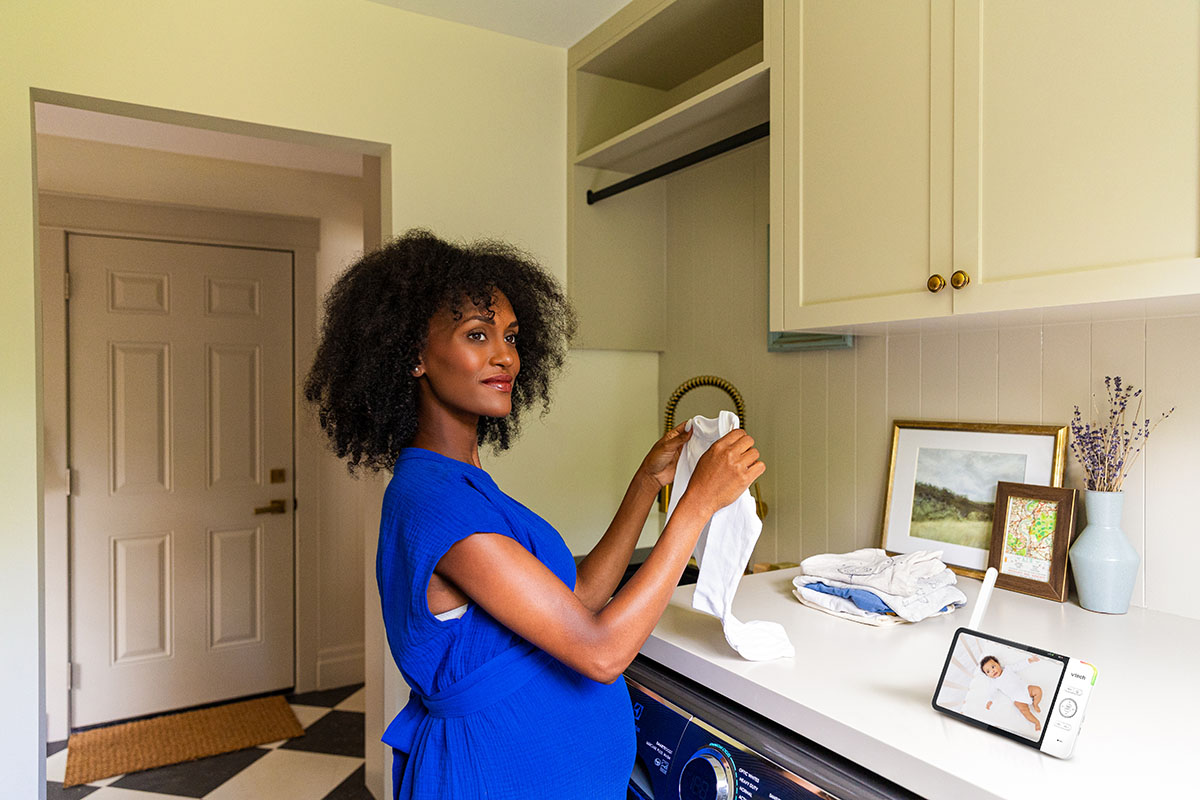 A woman in a blue dress folds laundry in a well-lit room with a photo frame, vase, and tablet—beside her, massage guns rest on the counter, ready to soothe tired muscles after chores.