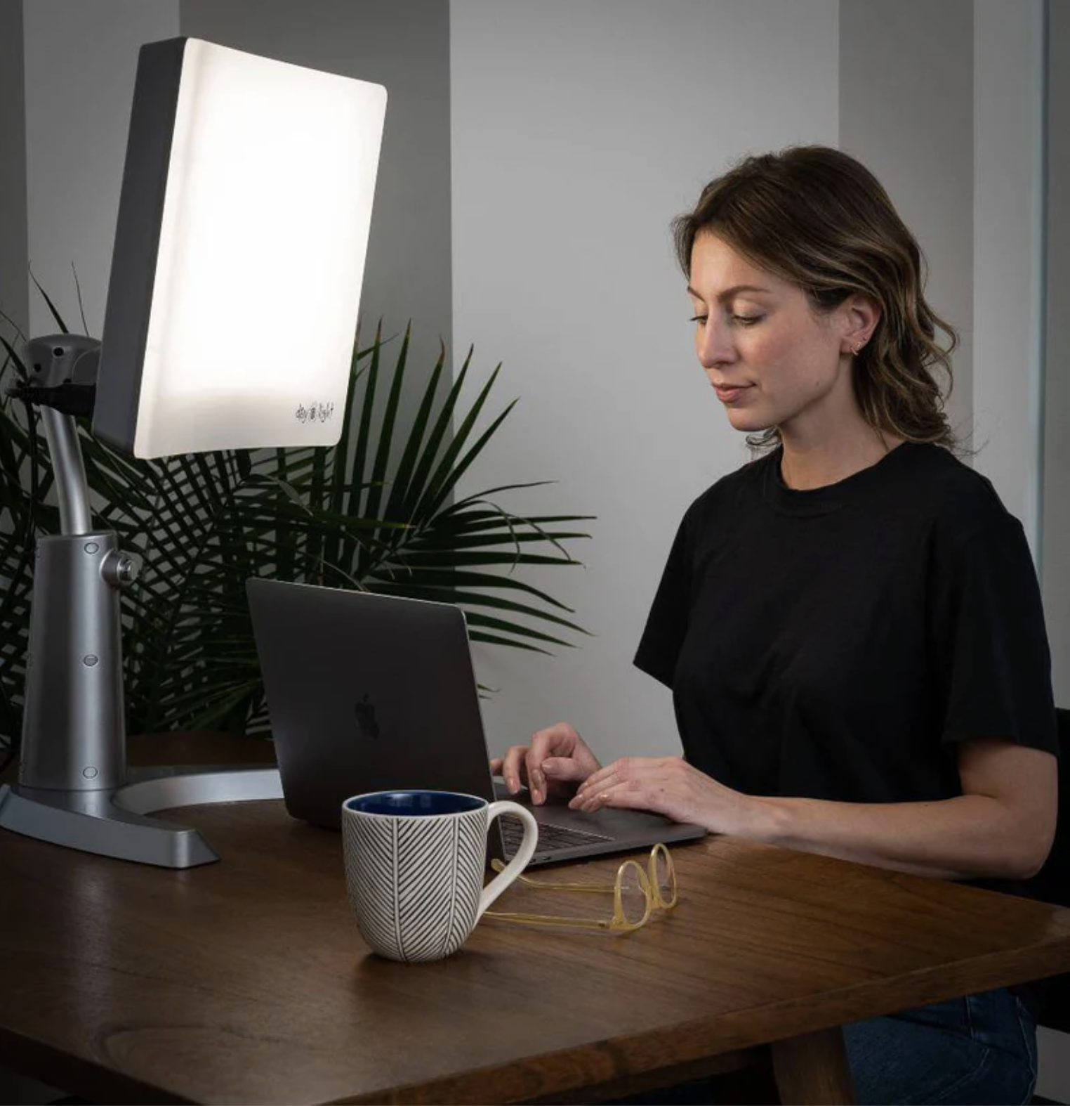 A woman sits at a desk using a laptop with a large light therapy lamp nearby. On the wooden desk are a mug, a pair of glasses, a plant, and a massage gun, suggesting a focus on both productivity and well-being.