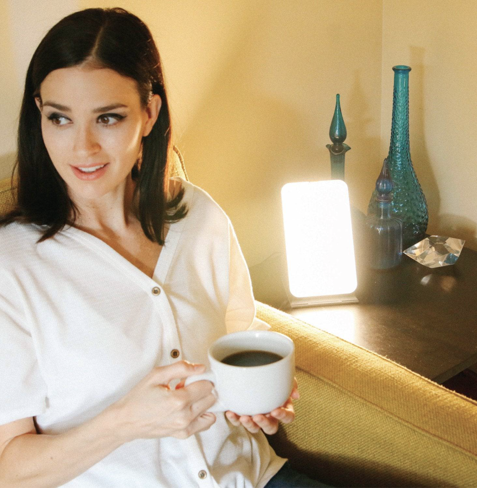 A woman sits on a yellow chair holding a mug of coffee, with a massage gun on the table beside her, alongside a bright light therapy lamp and decorative items.
