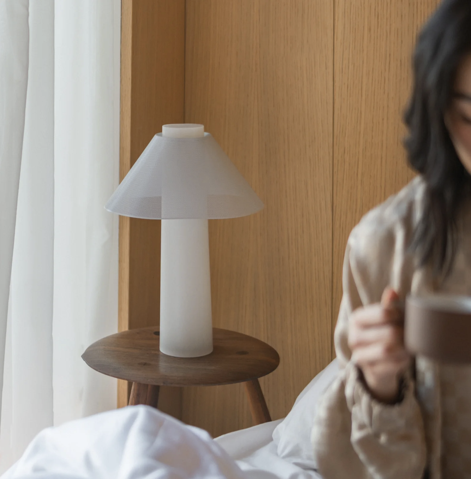 A white table lamp on a wooden bedside table next to a bed, with massage guns neatly placed alongside and a person holding a cup partially visible in the foreground.