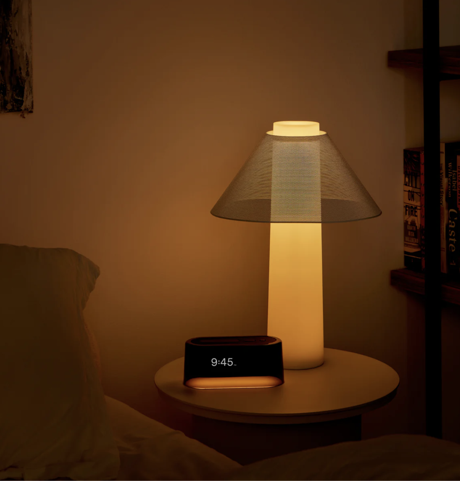A bedside table with a glowing lamp, massage guns, and a digital clock displaying 9:45, next to a bed and bookshelf in a dimly lit room.