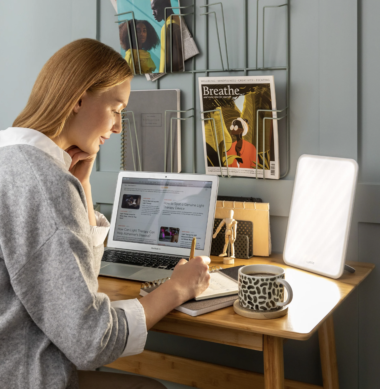 A woman sits at a desk using a laptop, writing in a notebook, with a mug, an illuminated light therapy lamp, and massage guns nearby. Magazines are displayed on the wall behind her.