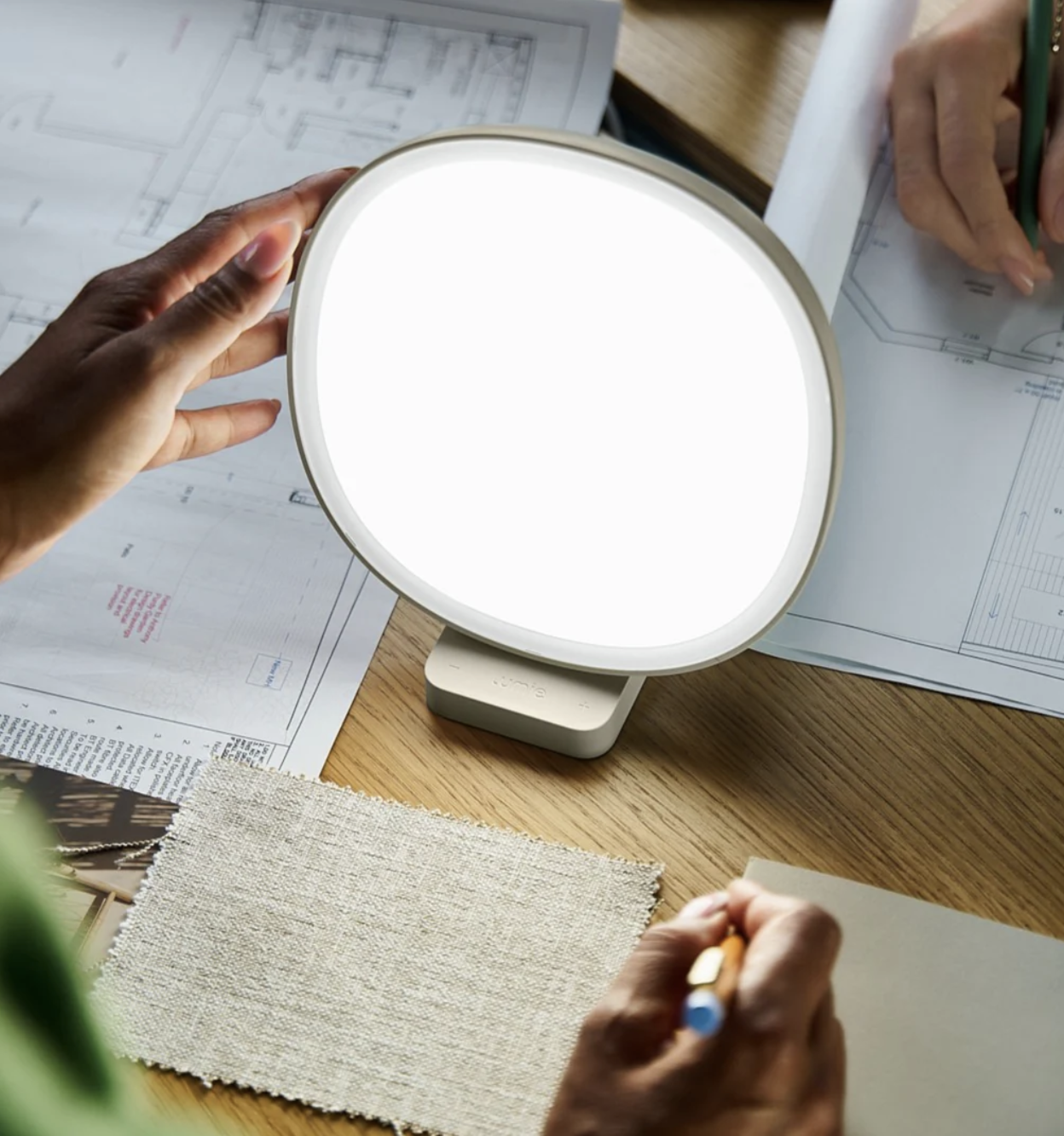 Two people work at a table with architectural plans, fabric swatches, massage guns, and a bright light therapy lamp in the center.