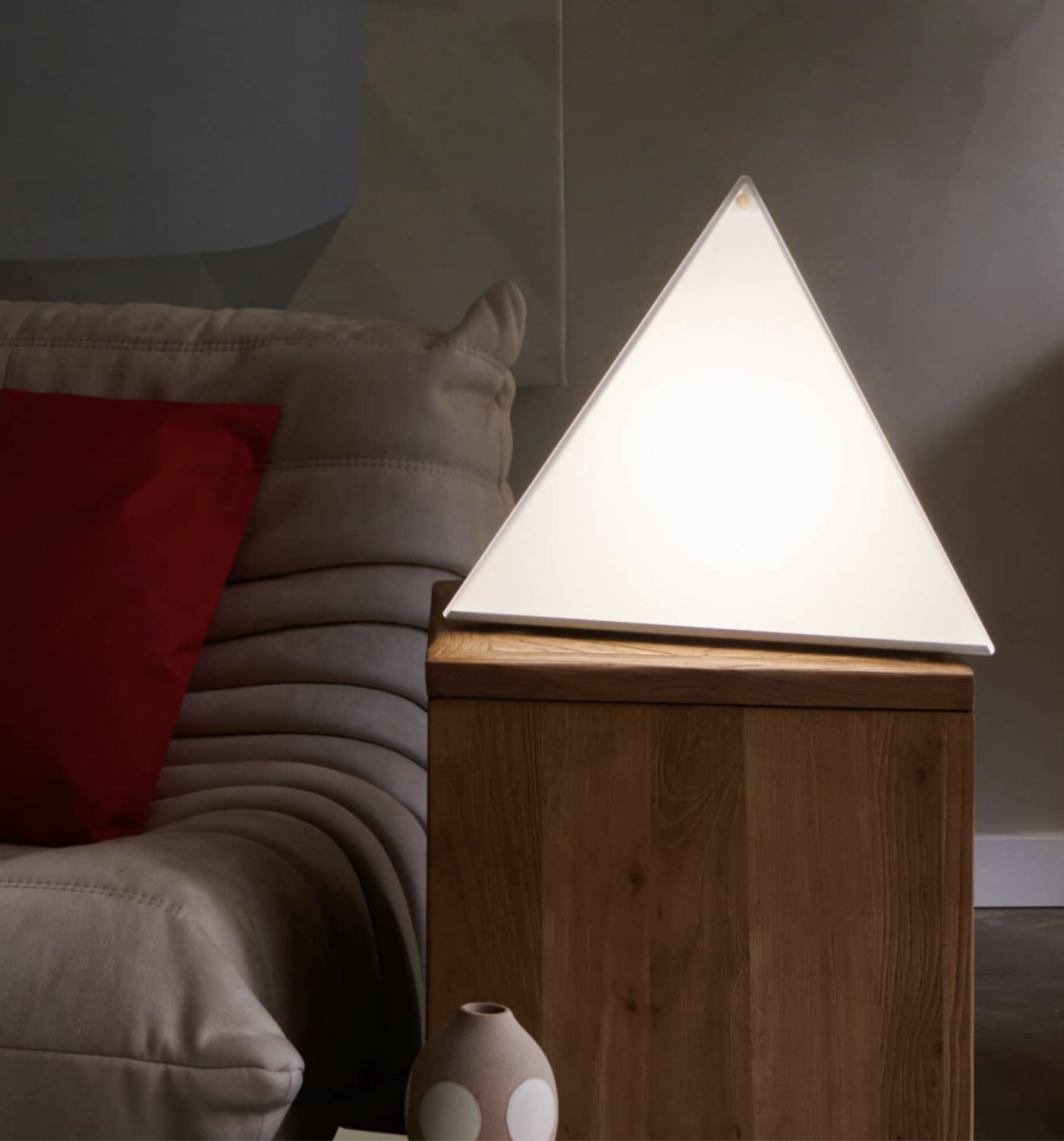 A triangular lamp sits on a wooden side table next to a beige couch with a red pillow. A small decorative vase and a sleek massage gun are also arranged on the table.