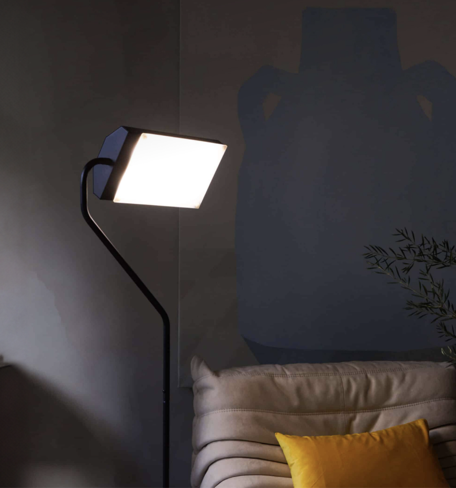 A modern floor lamp with a square shade glows beside a light-colored sofa with a yellow cushion, set against a dark wall with a large abstract design—perfect for relaxing with massage guns after a long day.