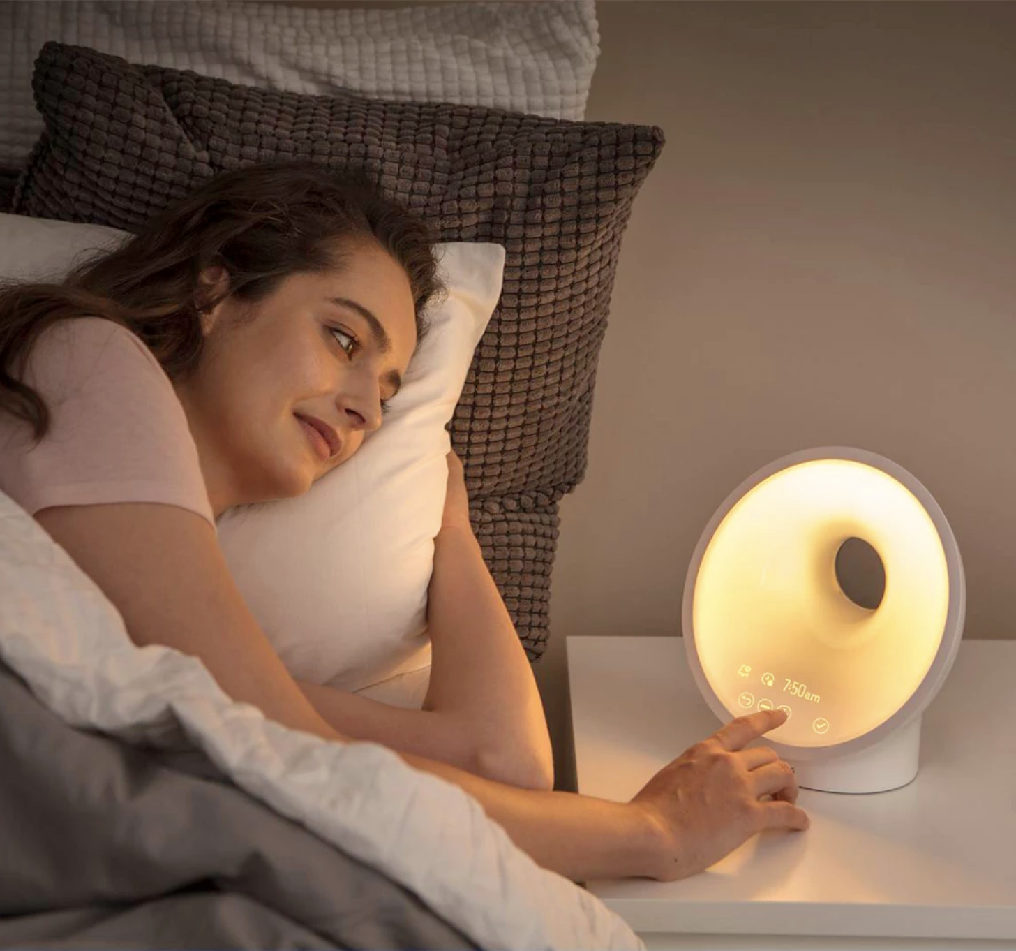 A person lying in bed touches a light therapy alarm clock on a bedside table, which emits a warm yellow glow; next to it, massage guns rest within easy reach for a soothing start to the day.