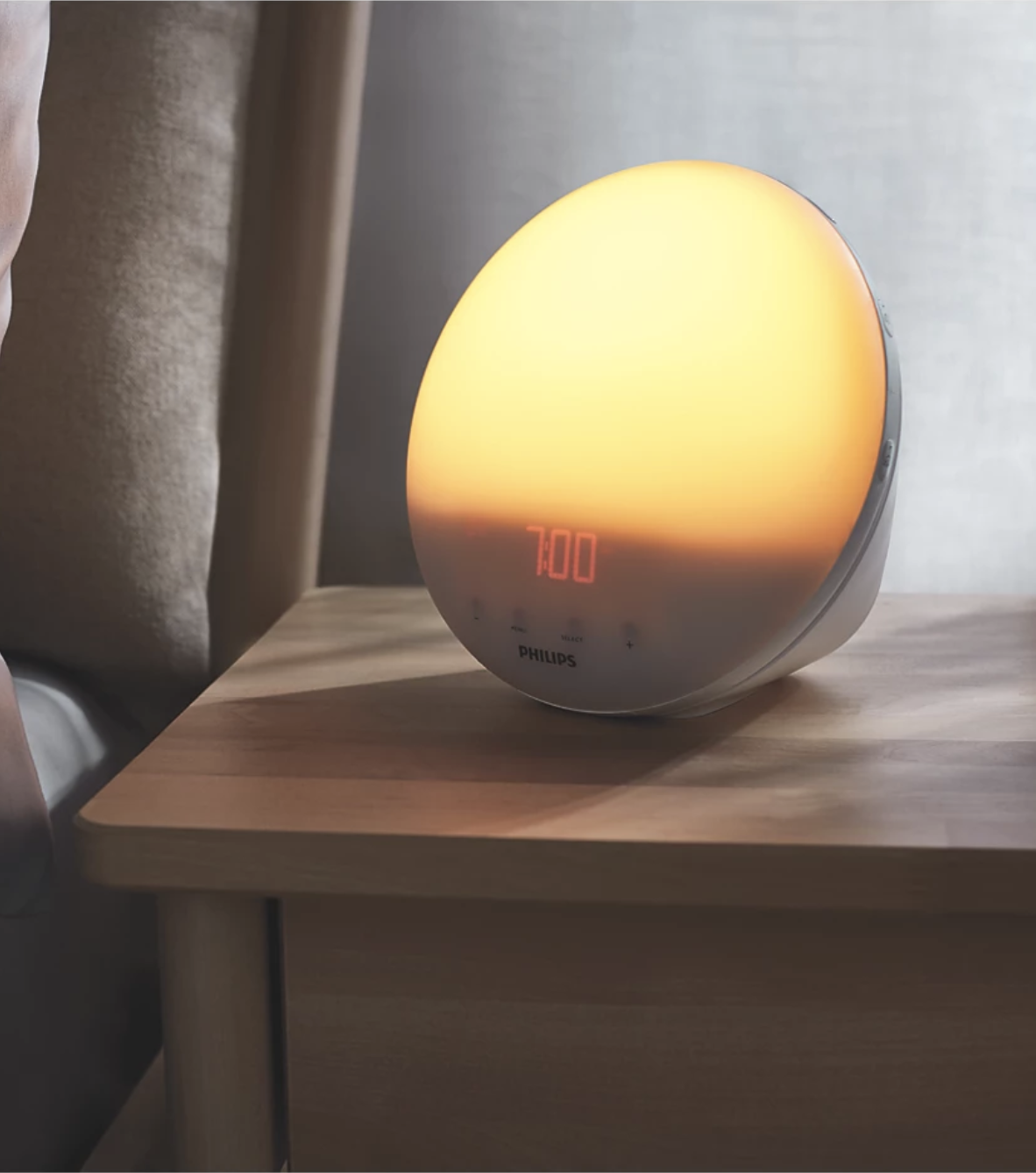 A Philips sunrise alarm clock with an orange-yellow glow displays 7:00 on a wooden bedside table next to a bed and a pair of massage guns, ready for a soothing start to the day.
