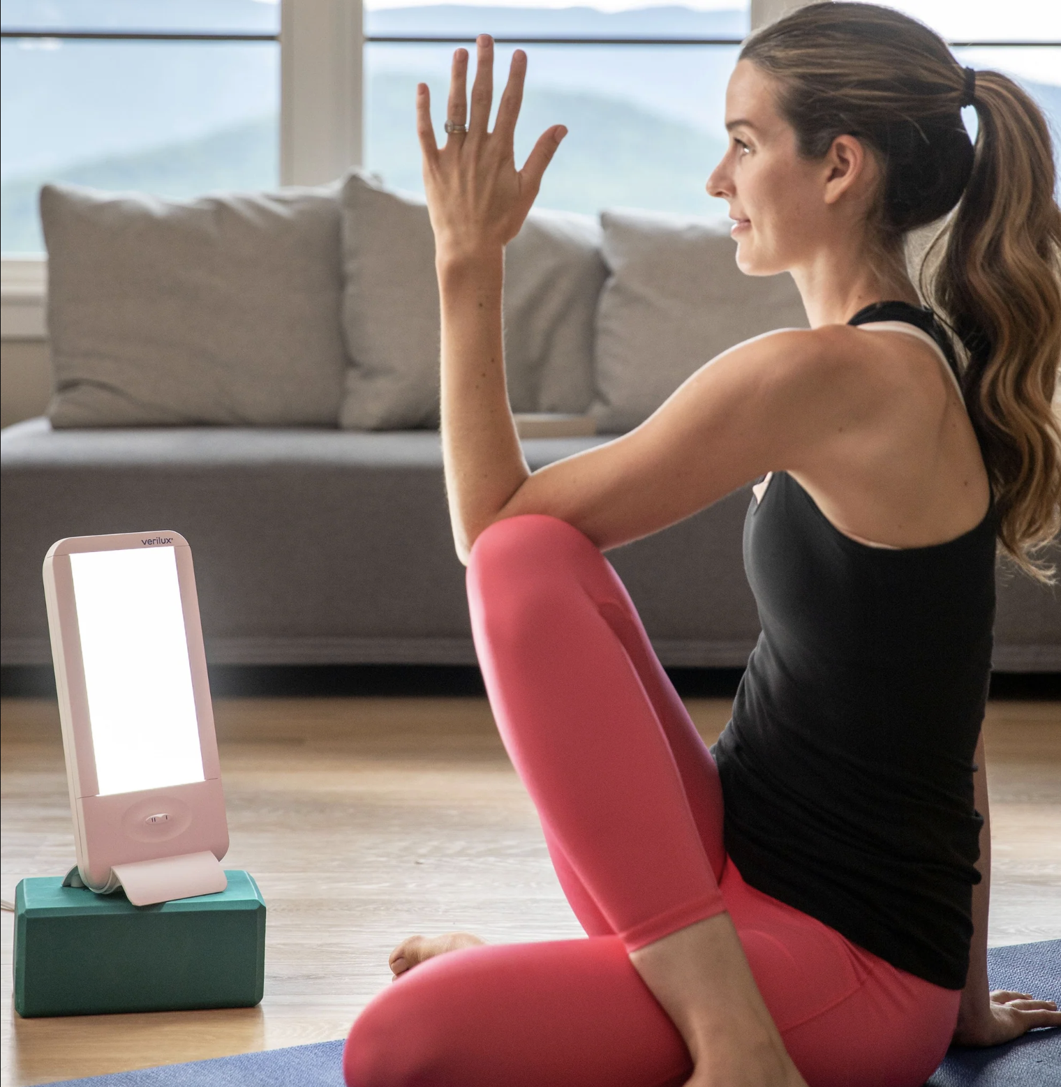 A woman in workout clothes sits on a yoga mat indoors, facing a bright therapy light placed on a small block, while massage guns rest nearby for post-workout recovery.