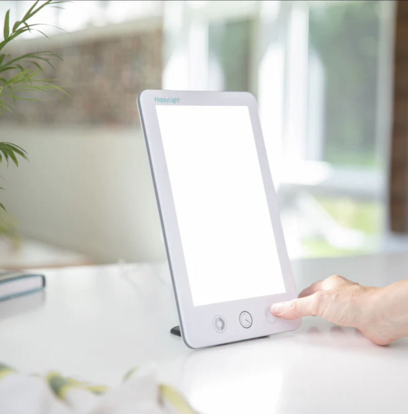 A hand presses a button on a rectangular light therapy lamp, placed on a white surface near a window alongside massage guns.