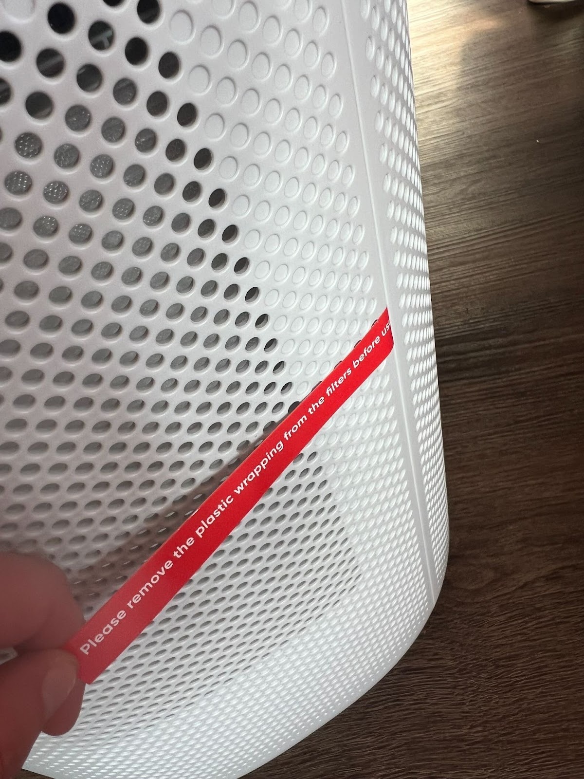 A hand holds a red label on a white air purifier that reads, "Please remove the plastic wrapping from the filter before use.