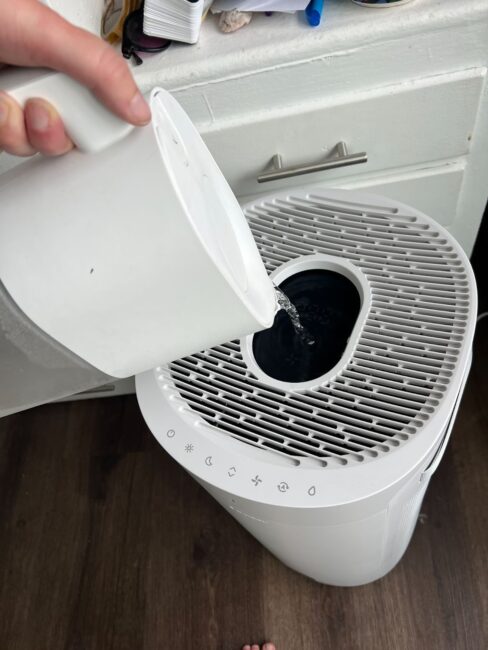 A person pours water from a white pitcher into the top opening of a white humidifier placed on a dark floor.
