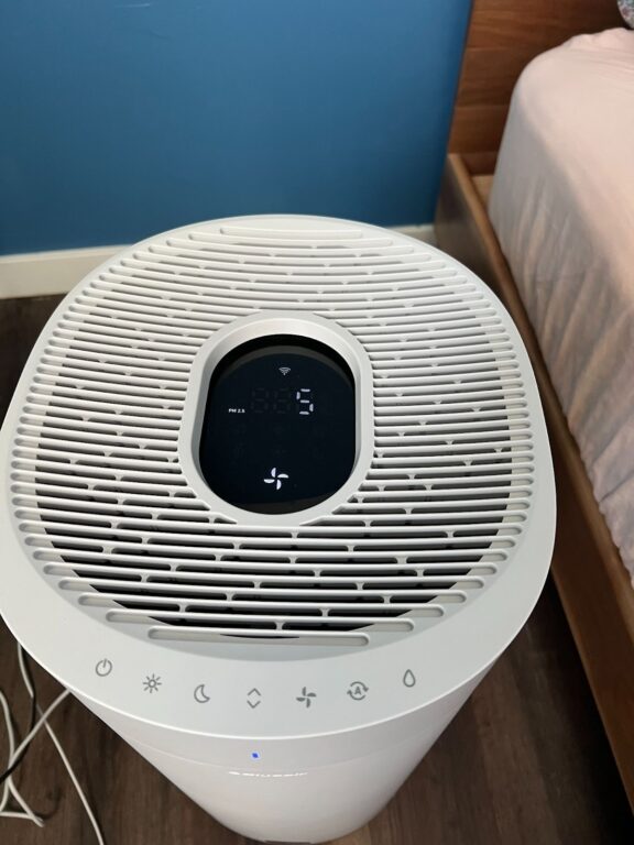 A white air purifier with a digital display and control panel sits on a wooden floor next to a bed with a pink blanket.