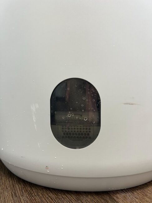 Close-up of a white appliance with a transparent oval window showing some condensation and internal components, placed on a wooden surface.