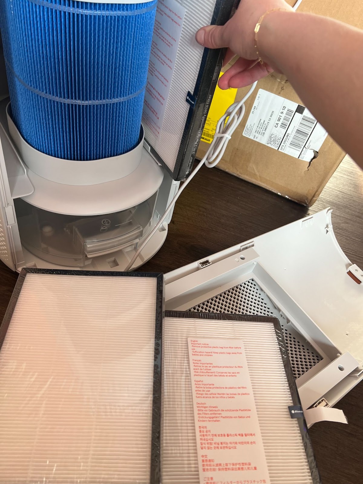 A person replaces filters in an air purifier, with new filters, packaging, and an open box visible on a wooden surface.
