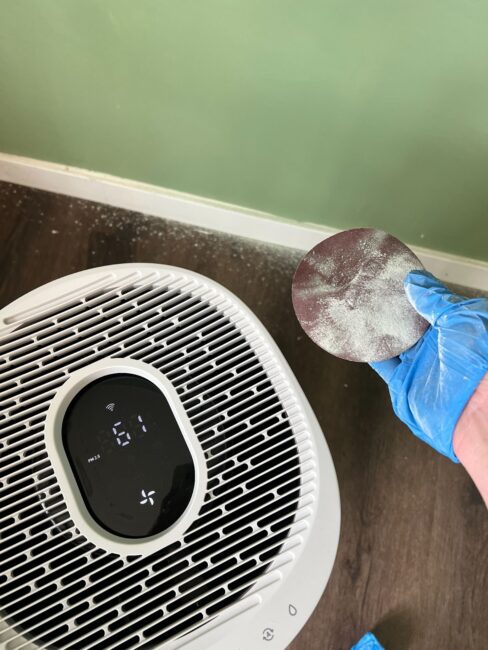 A gloved hand holds a dusty sanding disc near an air purifier on a wooden floor, with white dust scattered beside a green wall.
