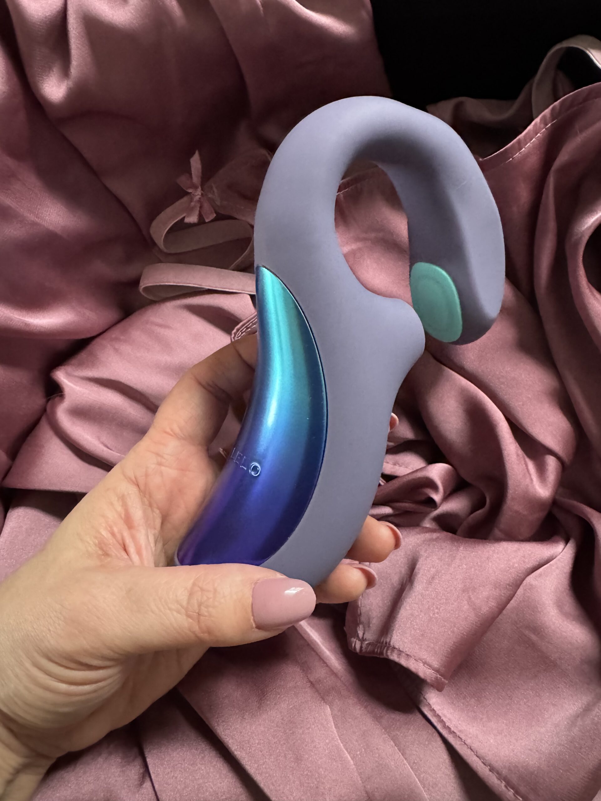 A hand holds a curved, dual-toned gray and blue personal massager against a background of pink satin fabric.