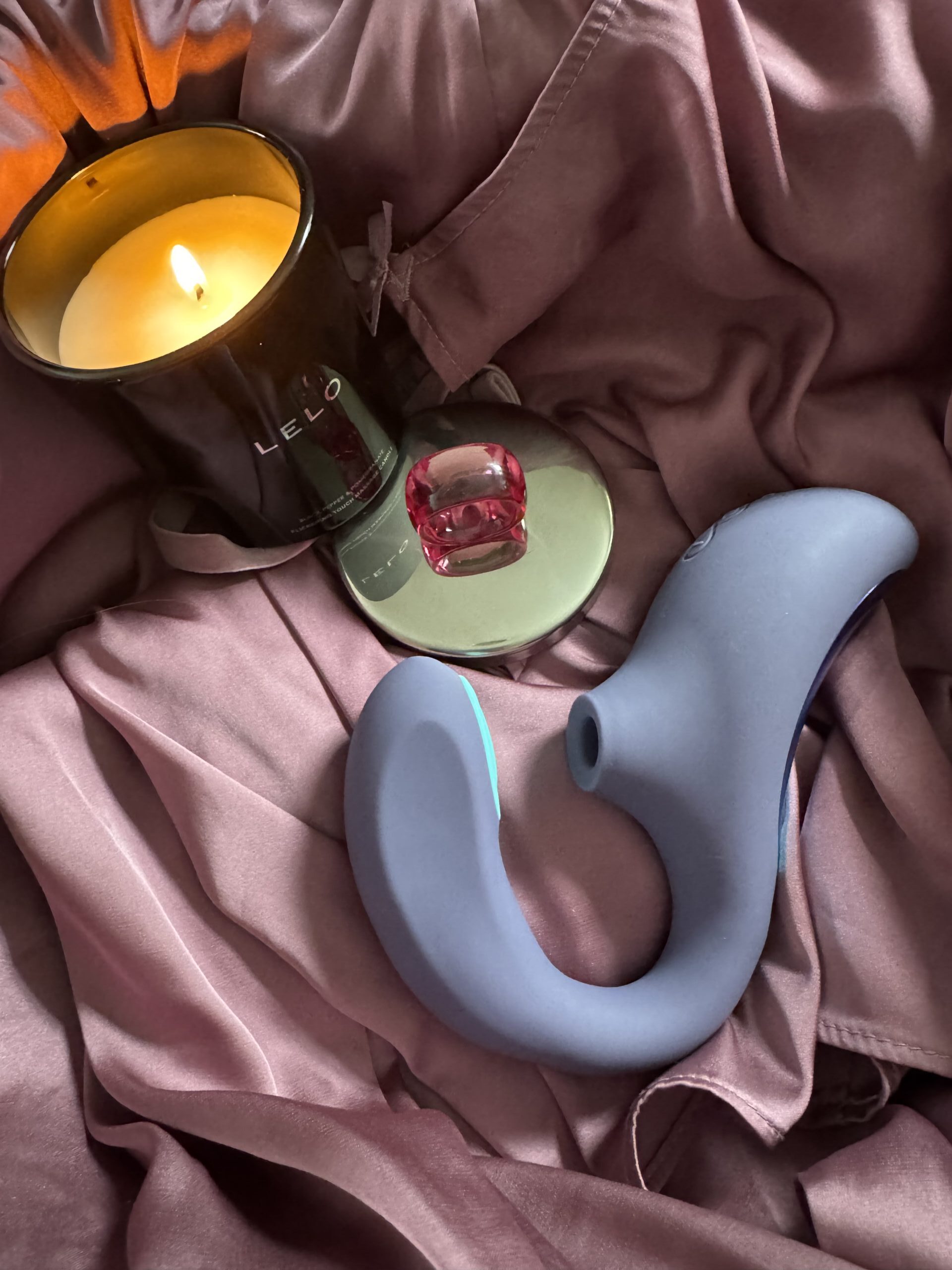 A grey silicone sex toy, a pink ring, a lit black candle, and a small mirror on mauve-colored fabric.