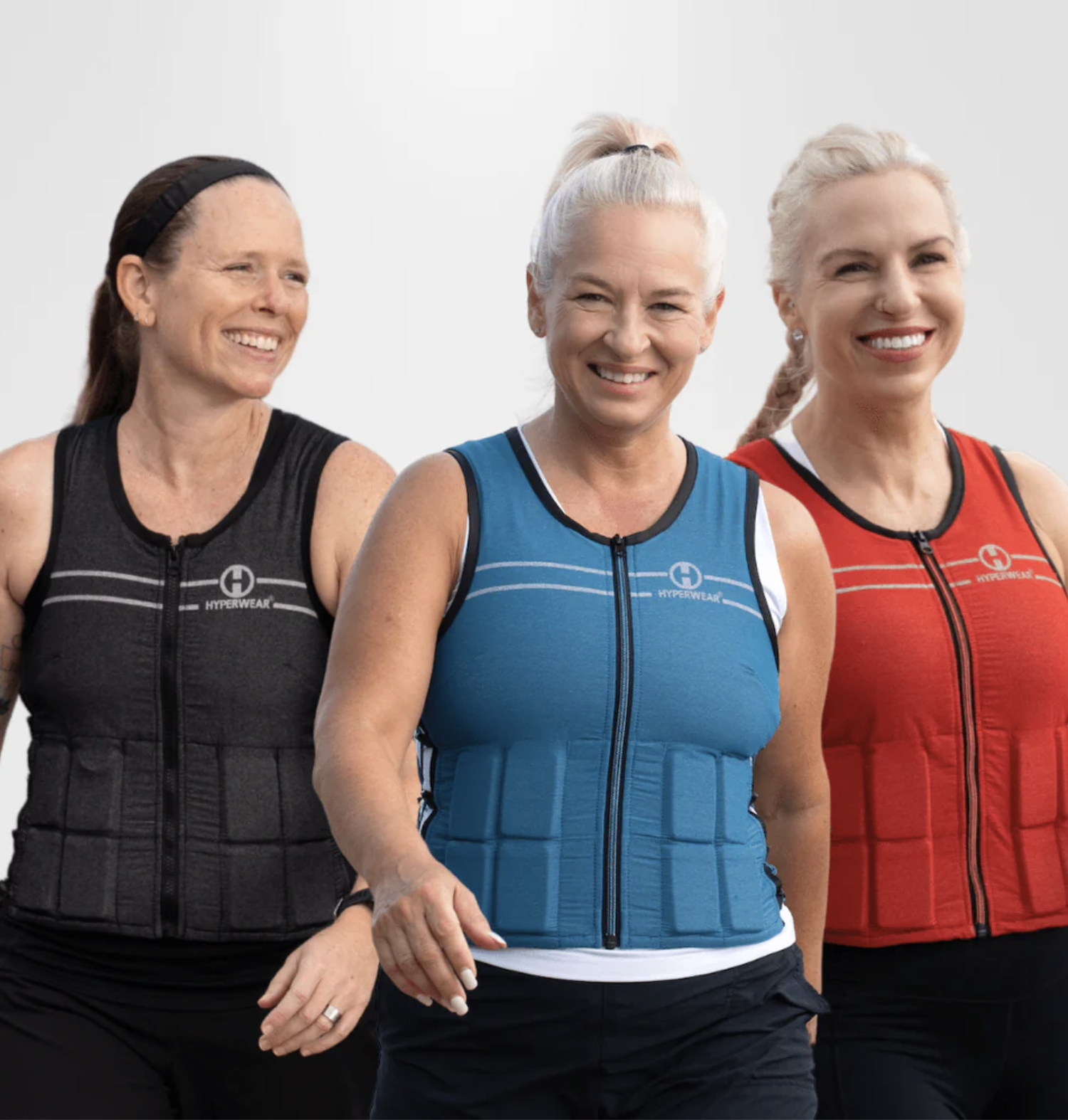 Three women in athletic clothing and weighted vests—black, blue, and red—walk together, smiling against a plain background, likely post-workout and ready to relax with their massage guns.