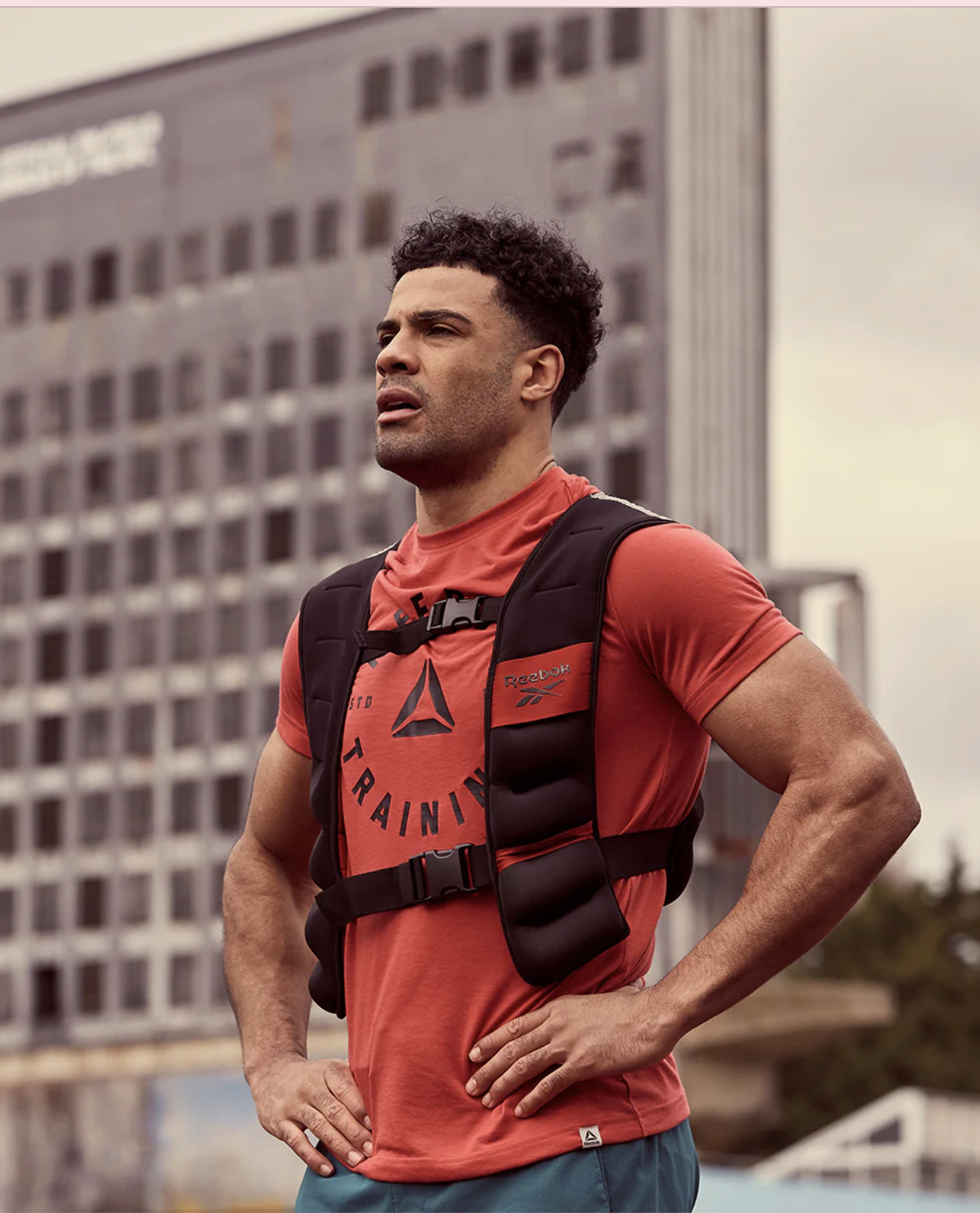 A man in a red Reebok t-shirt and weighted vest stands outdoors with his hands on his hips, likely ready to recover after his workout with massage guns; a high-rise building rises in the background.