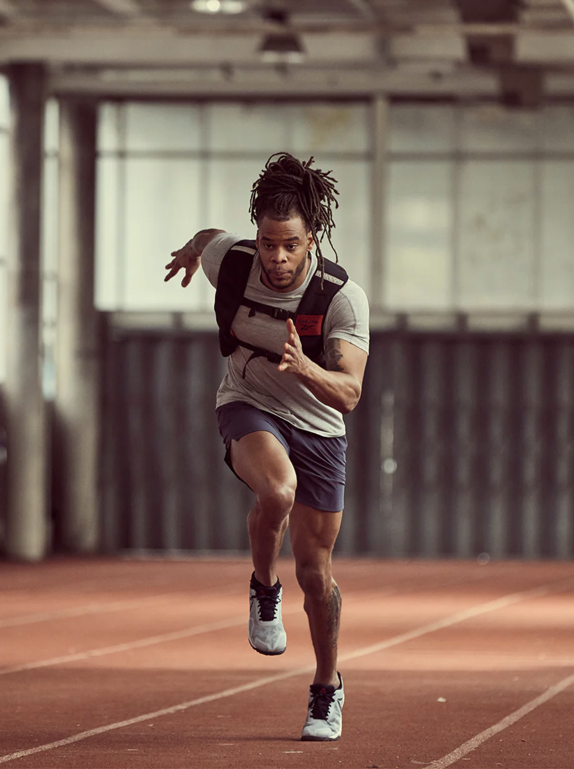 A man wearing a weighted vest sprints on an indoor track, focused and mid-stride, with blurred background details—his recovery made easier with massage guns after intense training sessions.