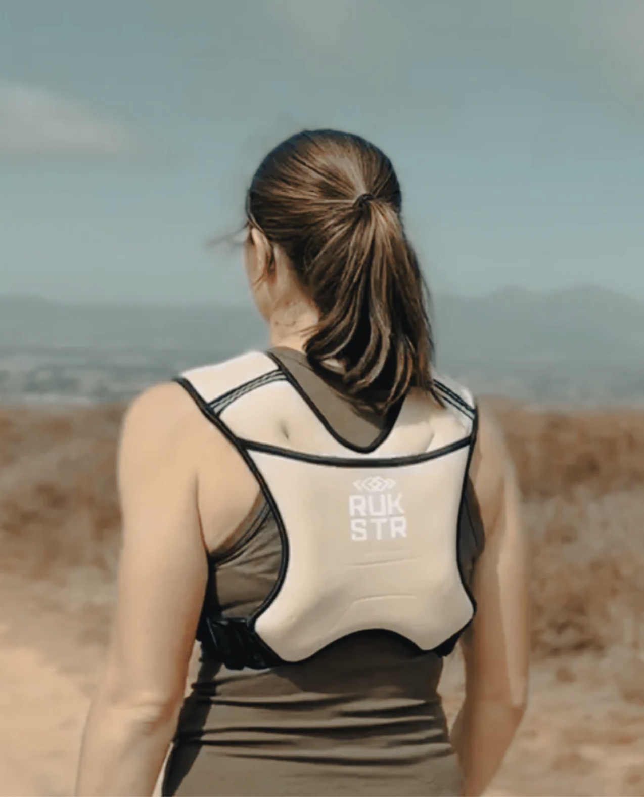 A woman wearing a white RUK STR posture corrector stands outdoors, facing away from the camera with a scenic landscape in the background, showing how posture support pairs perfectly with recovery tools like massage guns.