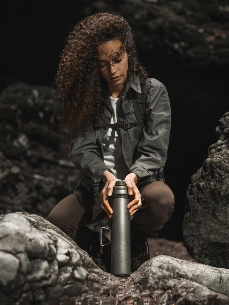 A person with curly hair sits on a rock in an outdoor setting, holding a smart waterbottle and looking down at it, surrounded by trees and rocks in the background.