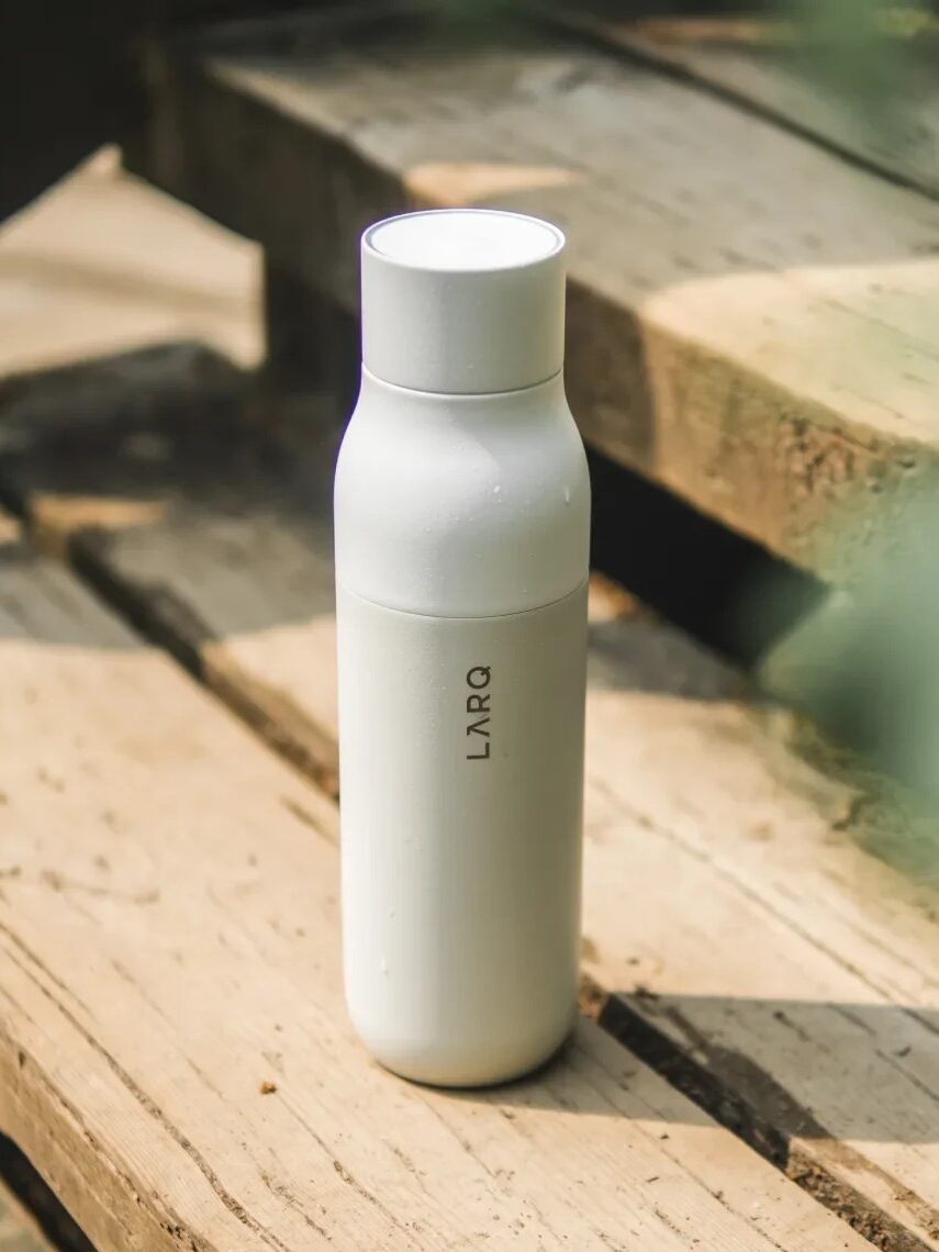A white LARQ smart waterbottle is placed upright on a weathered wooden surface outdoors, with sunlight casting soft shadows.