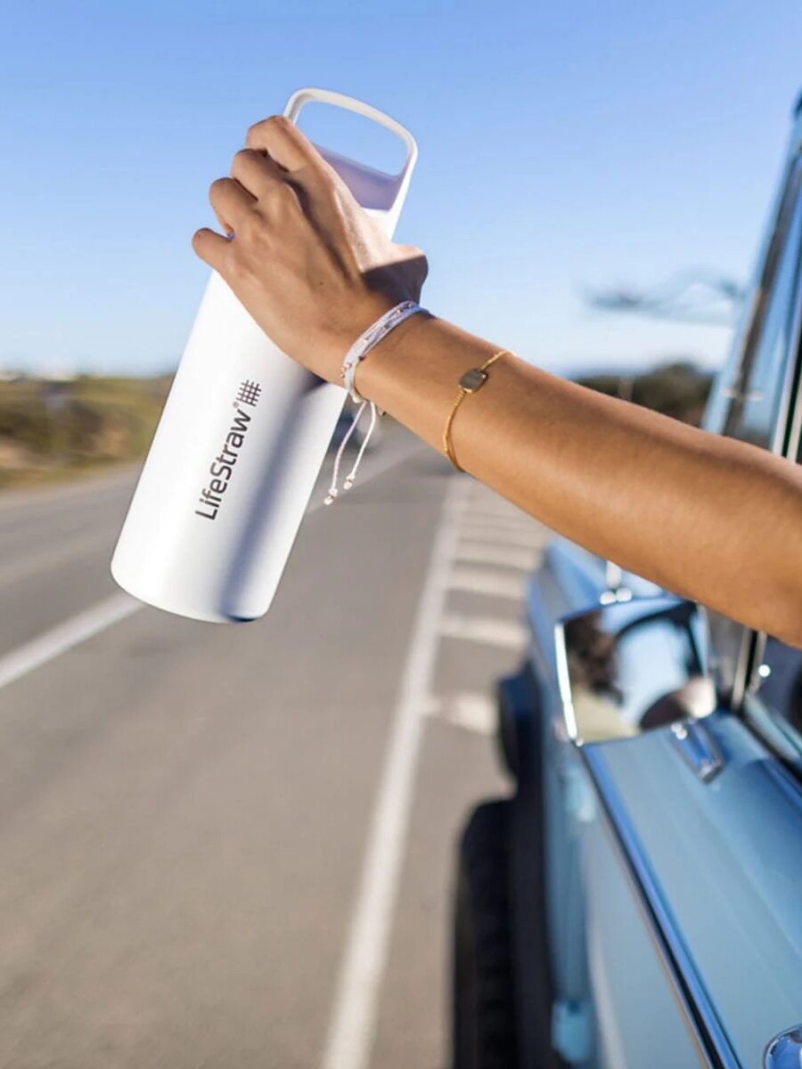 A person holds a white LifeStraw smart waterbottle out of the window of a blue vehicle parked on the side of a road.