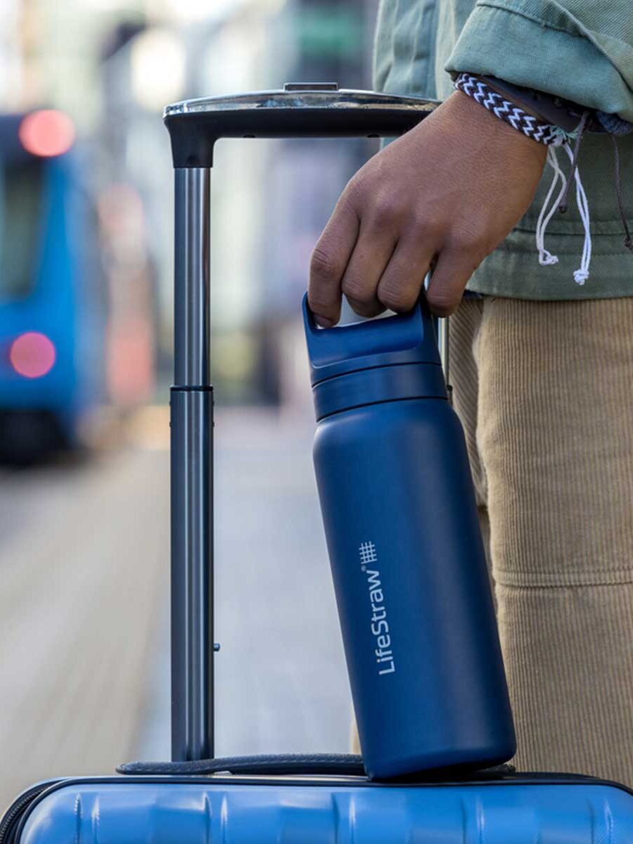 A person holds a blue smart waterbottle by the handle while gripping the handle of a blue suitcase on a city sidewalk.